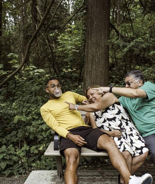 A man in a yellow shirt blows a bubblegum bubble while sitting on a forest park bench as an older couple beside him laugh and reach across playfully to try to pop it.