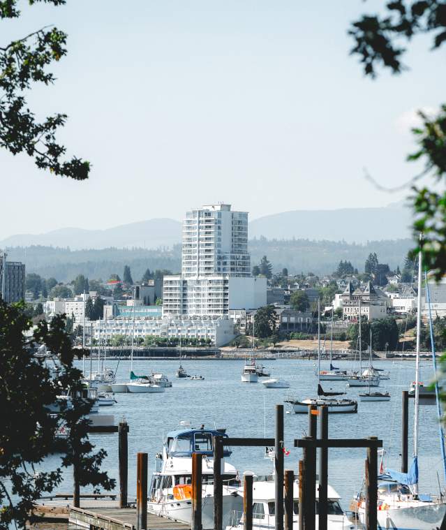 A view of Nanaimo’s waterfront with anchored boats and marina in the foreground, city skyline and distant hills visible through tree branches.