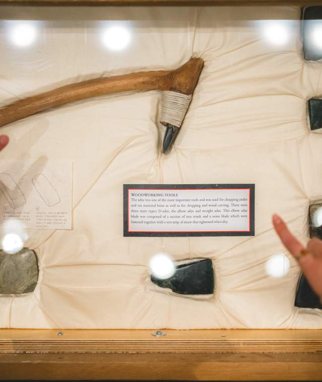 Two people point at a museum display featuring traditional woodworking tools and small stone adzes arranged behind glass.