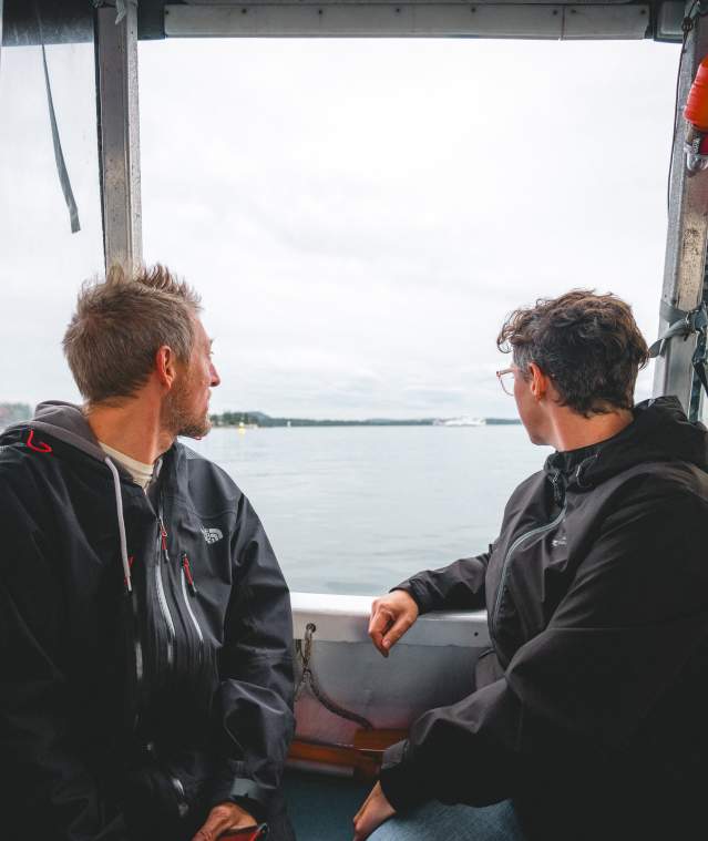 Two individuals sit inside a small boat, looking out over calm coastal waters toward a distant shoreline under an overcast sky.