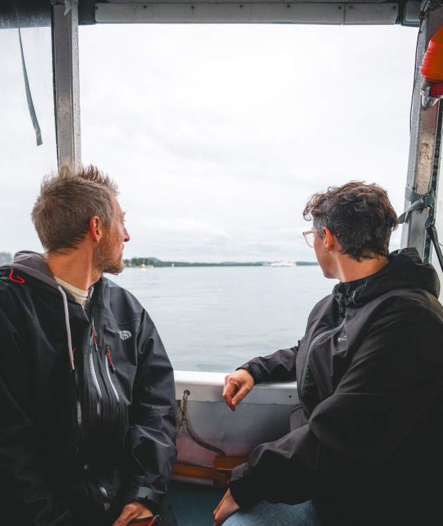 Two individuals sit inside a small boat, looking out over calm coastal waters toward a distant shoreline under an overcast sky.
