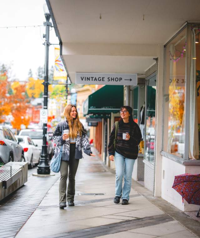 Two people stroll past shops on a rainy autumn day in downtown Nanaimo, holding coffee as colourful fall trees line the street.