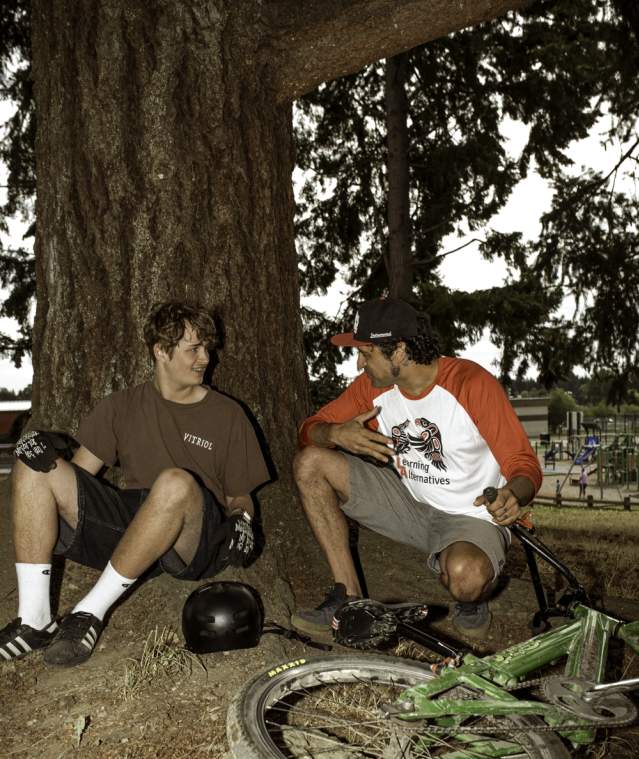 Two men sitting leaning on a tree with a bike.