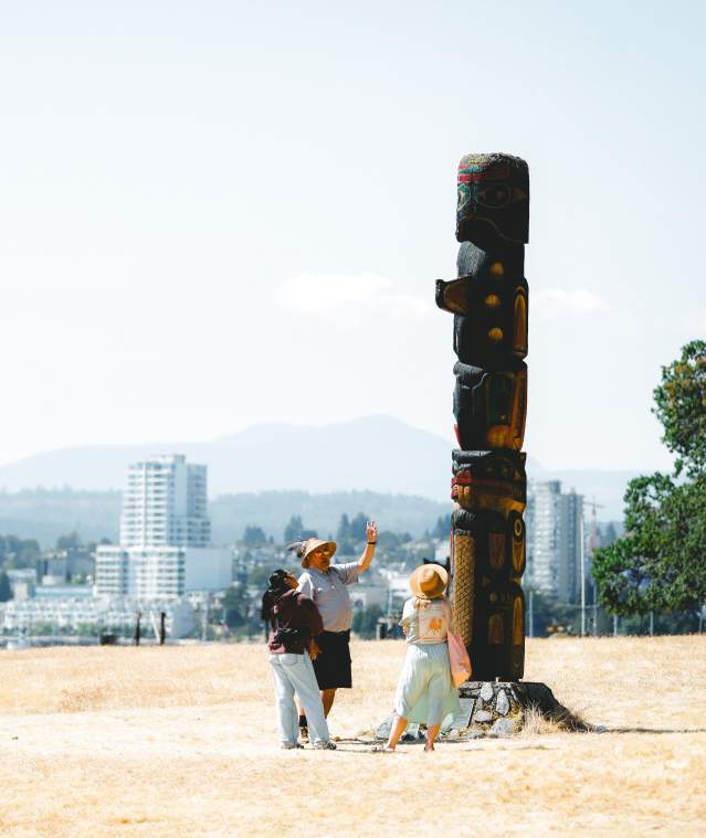 Three people stand at the base of a totem pole on Saysutshun with Nanaimo’s skyline visible across the harbour in the background.