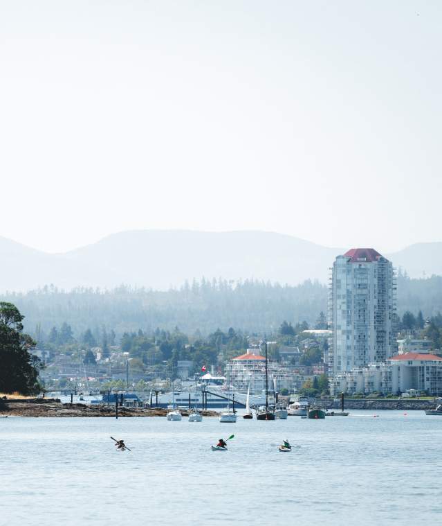 Three kayakers paddle across calm water with a forested island on one side and Nanaimo’s skyline in the distance.