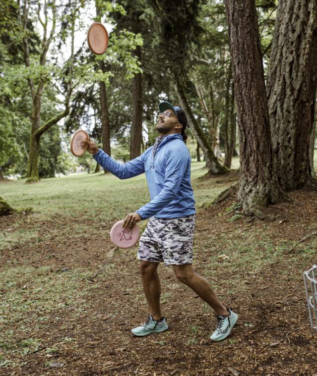 A person plays disc golf in a wooded park, tossing a disc toward a metal basket surrounded by trees.