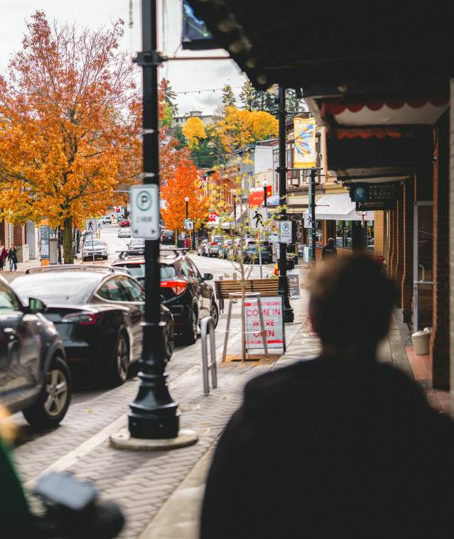 People walk along a downtown Nanaimo street with shops, parked cars, and vibrant autumn trees lining the road.