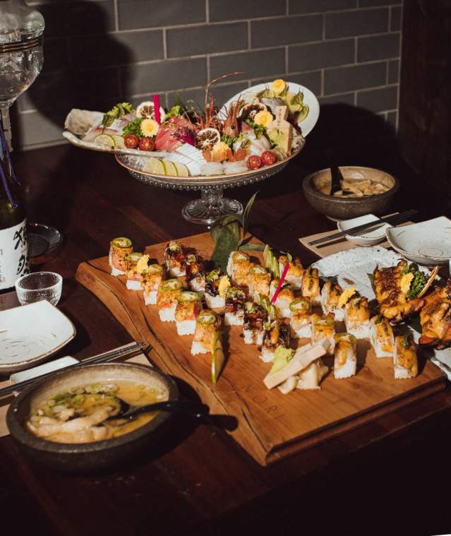 A restaurant table filled with sushi rolls, sashimi, soup, and drinks arranged on wooden boards and serving dishes.