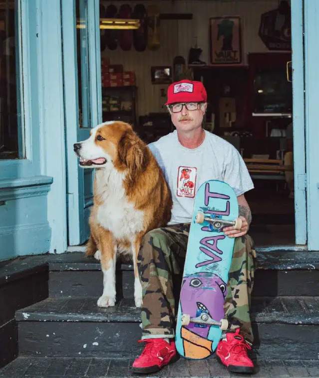 Aa man and his dog sitting with a skateboard.