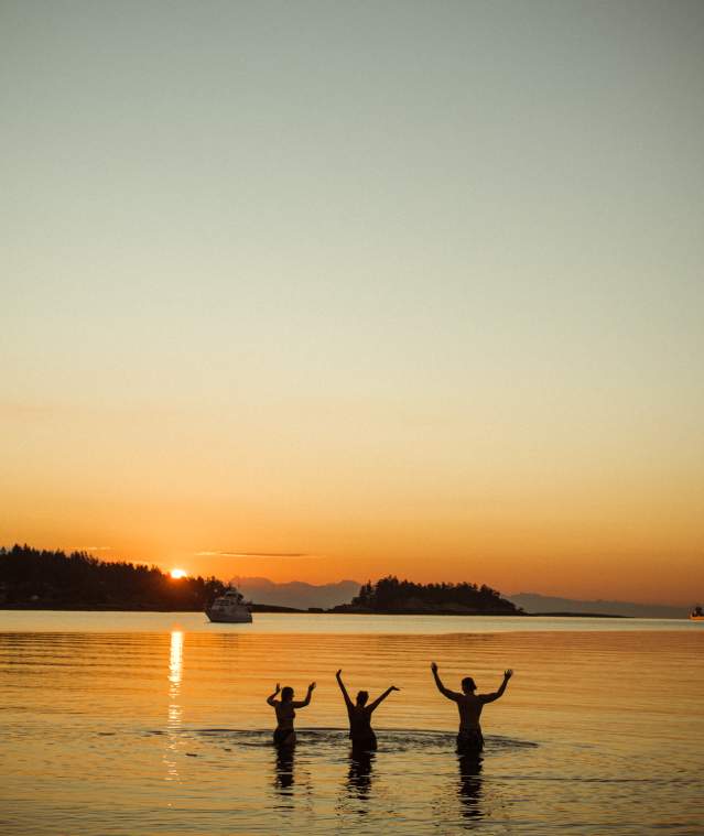 Three people standing in calm ocean water at sunset with arms raised.