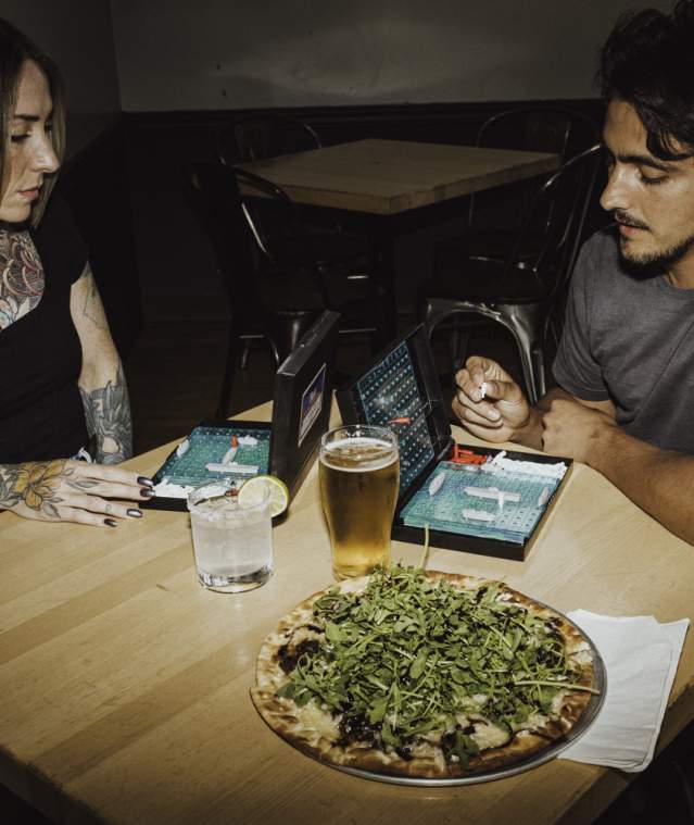 Two people play a tabletop game at a restaurant table with pizza and drinks in front of them.