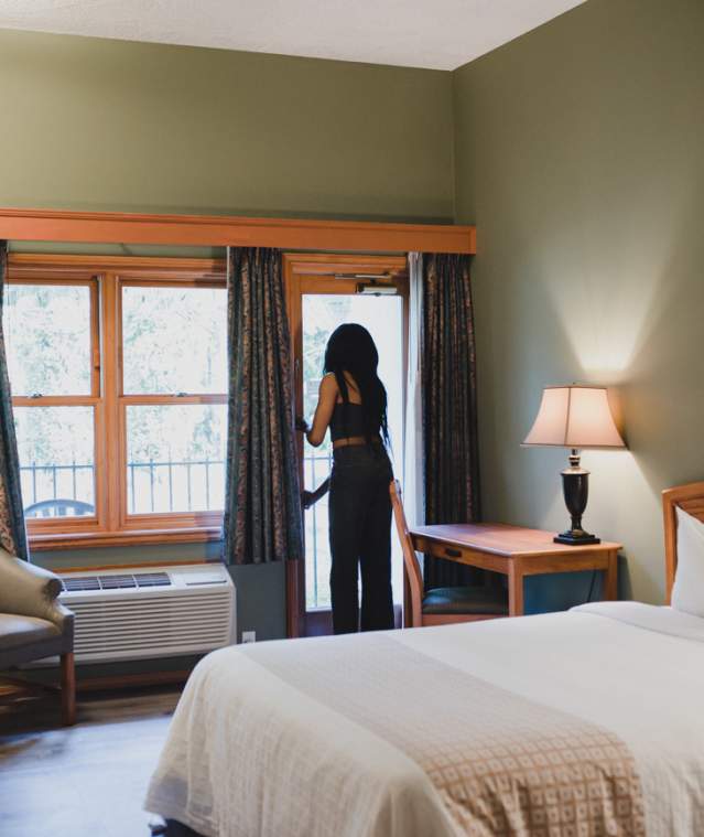 A cozy hotel room with a bed, desk, and seating area as a person opens a balcony door to let in natural light.