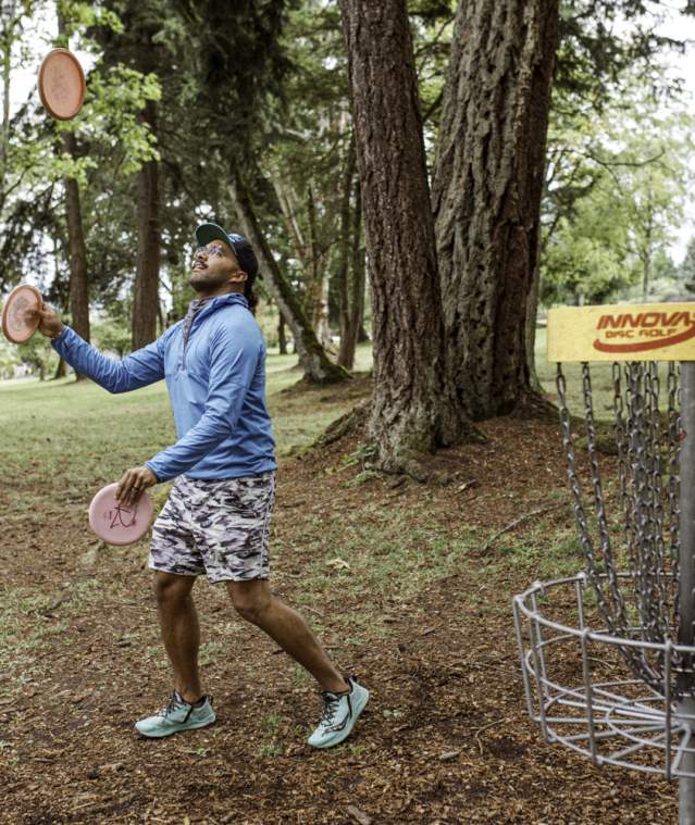 A person plays disc golf in a wooded park, tossing a disc toward a metal basket surrounded by trees.