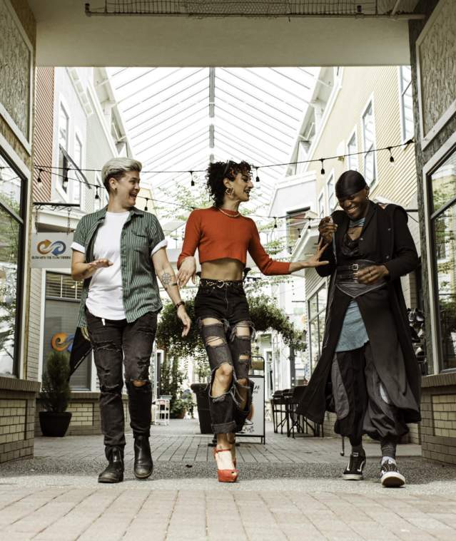 Three friends walk side by side, laughing in a bright, covered shopping walkway lined with storefronts.