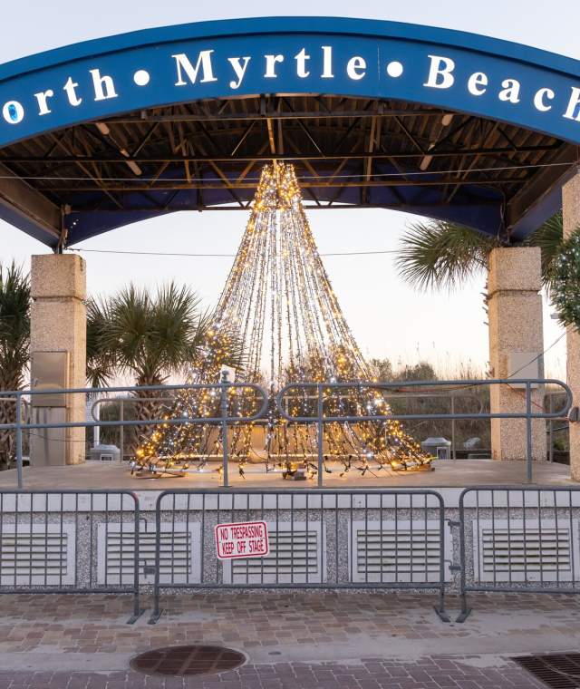 Holiday-themed stage at North Myrtle Beach with a conical Christmas light display in the center, flanked by decorated wreaths with red bows on granite columns. A blue arch above reads 'North Myrtle Beach.'