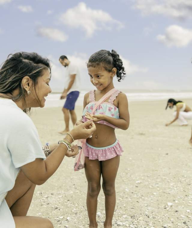 A woman showing seashells to a young girl on the beach, with other family members nearby exploring the sand.