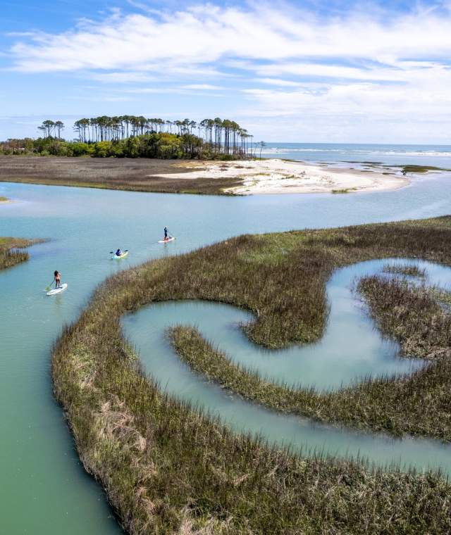 Aerial view of paddleboarders exploring winding salt marsh creeks near Waites Island in North Myrtle Beach, South Carolina, with pristine coastal scenery and clear blue skies.