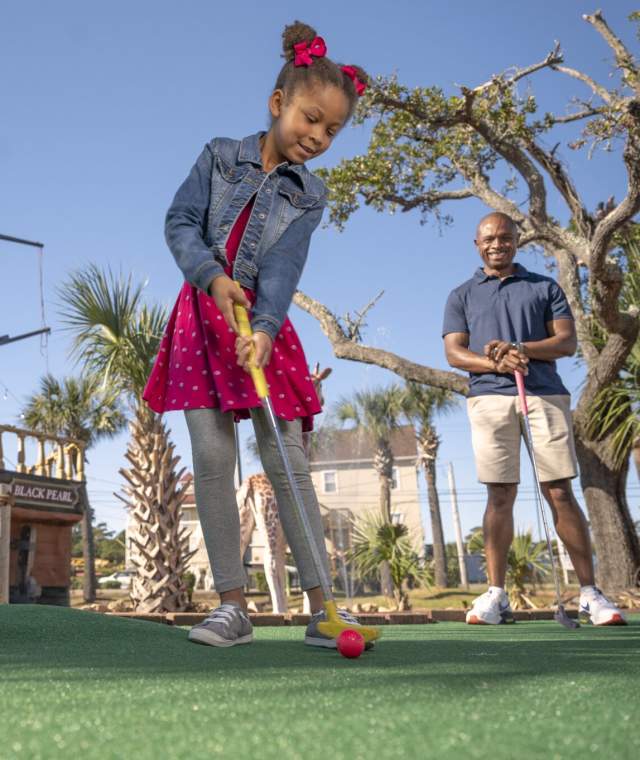Young girl playing miniature golf at a pirate-themed mini-golf course in North Myrtle Beach, South Carolina, with a smiling adult watching in the background under sunny skies and palm trees.