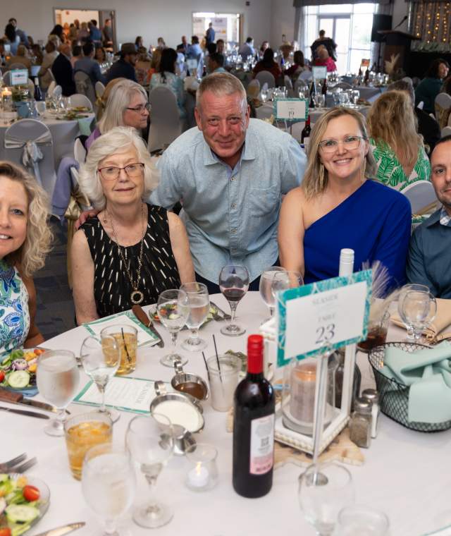 A group of five adults poses and smiles around a decorated dinner table at a formal event. The venue is filled with other seated guests, and the table is set with plates of salad, drinks, and a bottle of wine. A sign reading “NMB” is visible in the background, indicating a North Myrtle Beach gathering