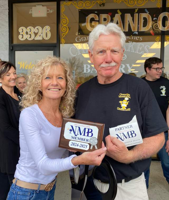 A smiling man and woman stand in front of Grandpa Moustache’s Cheesecakes holding Destination NMB Member and Partner plaques. They pose for the camera with others gathered behind them, and a “Grand Opening” banner is visible on the bakery window.