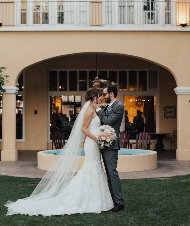 Groom in gray tuxedo kisses bride in mermaid-style wedding dress with veil on the forehead standing on the grass of the Crowne Plaza courtyard. A fountain can be seen in the background along with orange trees, and the historic, tan Crowne Plaza building with large archways.