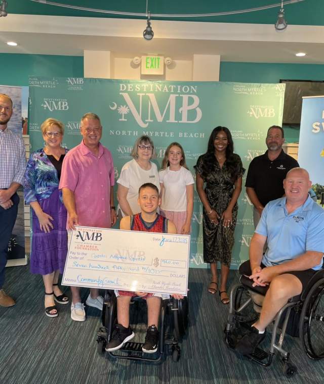 A group of people poses indoors in front of a “Destination NMB” backdrop, smiling and holding a large ceremonial check. Two individuals in wheelchairs are positioned at the front, emphasizing inclusivity. The check displays the North Myrtle Beach Chamber Foundation Logo.