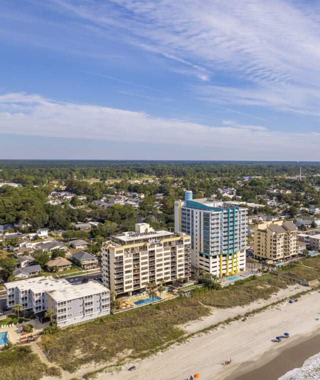 Aerial view of oceanfront hotels and vacation rentals along the sandy shoreline of North Myrtle Beach, South Carolina, with gentle waves, beachgoers, and a clear blue sky stretching over the coastal cityscape.