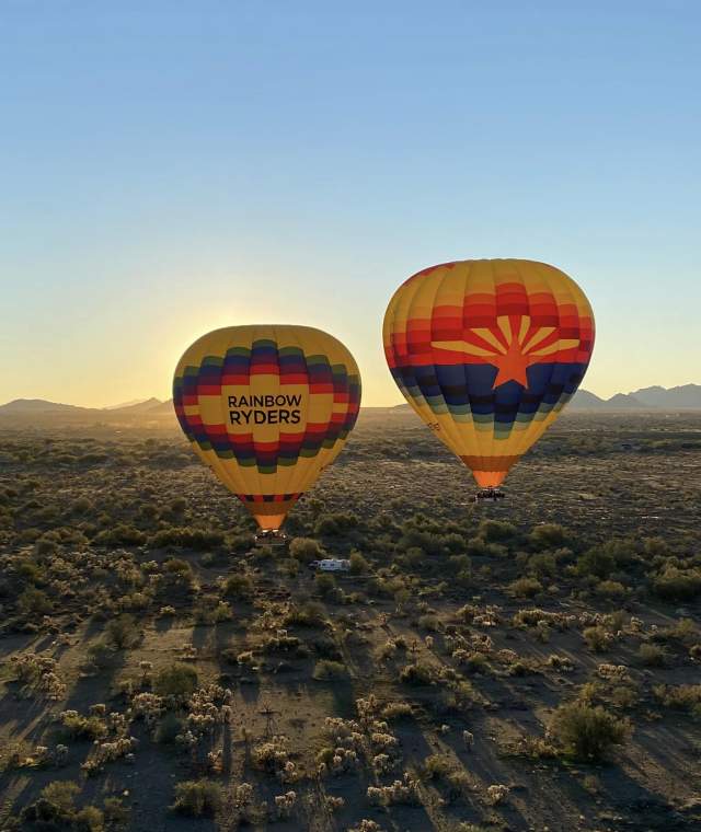 two hot air balloons, one with the name Rainbow Ryders on the front and the other with the Arizona state flag on the front, gliding over the mountainous desert