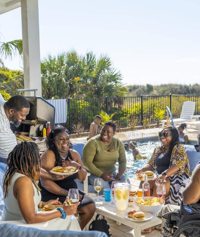 Multi-generational family enjoying a joyful reunion at a vacation rental in North Myrtle Beach, South Carolina, gathered around a poolside table with food and drinks, celebrating quality time together in a warm coastal setting.