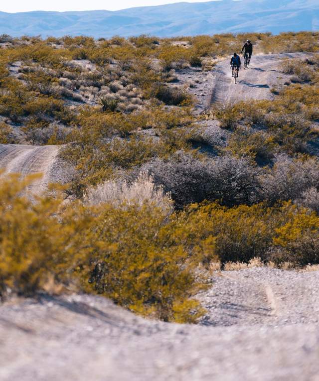 Two people riding a bicycle on a dusty, gravel trail through a sparse desert landscape.