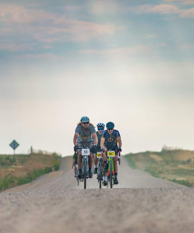A group of cyclists ride along a gravel road, surrounded by open fields and a hazy sky in Fort Collins