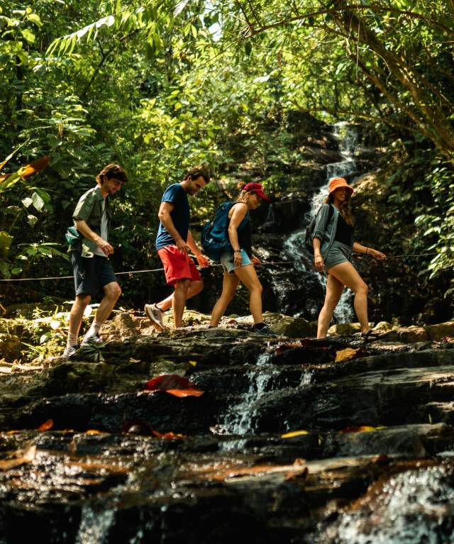 Un grupo de excursionistas cruzando un arroyo cerca de la cascada Pozo Azul, rodeado de muchos árboles y naturaleza.