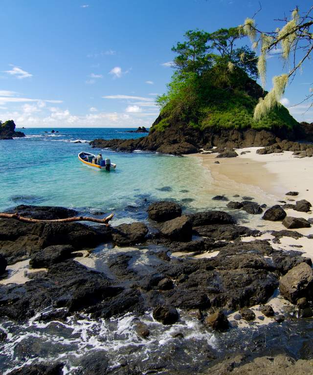 Pequeño barco navegando cerca de una costa rocosa en el Golfo de Chiriquí, con aguas turquesas cristalinas y verdes islas tropicales bajo un cielo azul claro.