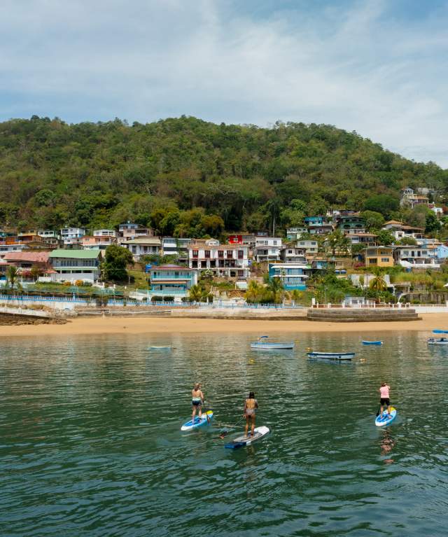 Three people practice paddleboarding in calm waters near the beach and colorful houses on the hillside of Taboga Island, surrounded by lush vegetation.