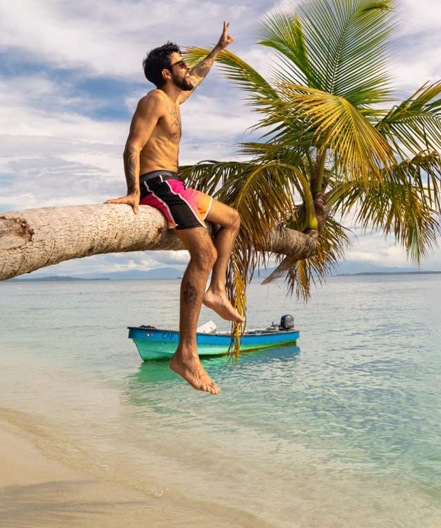 Man sitting on a palm tree on a caribbean beach with crystal clear light blue ocean and people in the background.