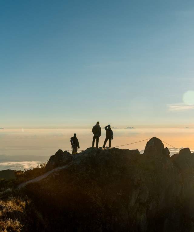 The image captures the silhouette of three people on the summit of a rugged mountain, likely Volcán Barú, at dawn or dusk, with the sun shining intensely to one side. Below them, a sea of clouds stretches to the horizon, creating a breathtaking landscape and an atmosphere of majestic tranquility.