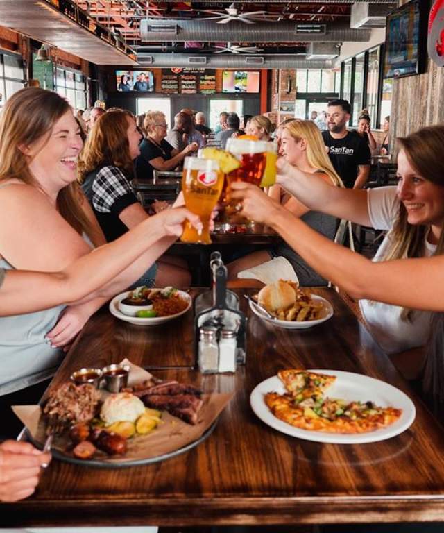 a busy restaurant interior in the background with focus on a table filled with plates of food and four ladies raising a glass of beer to cheer with
