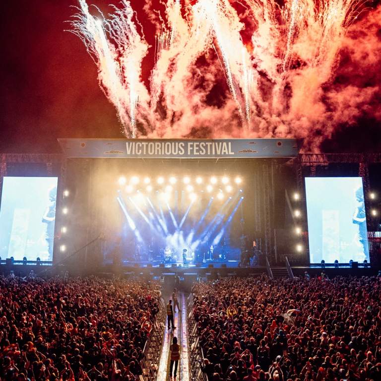 Photograph showing the culmination of a headline set at Victorious Festival, with red fireworks going off behind the stage and thousands of people gathered in front