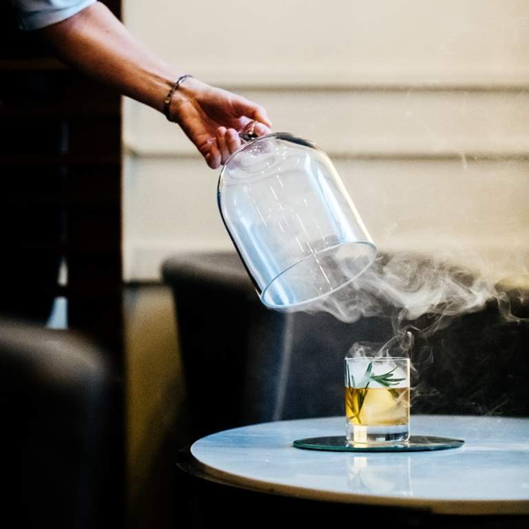 A waiter removing a glass cover that had been on a glass of whiskey to smoke it.  Smoke is still lingering in the air around the glass