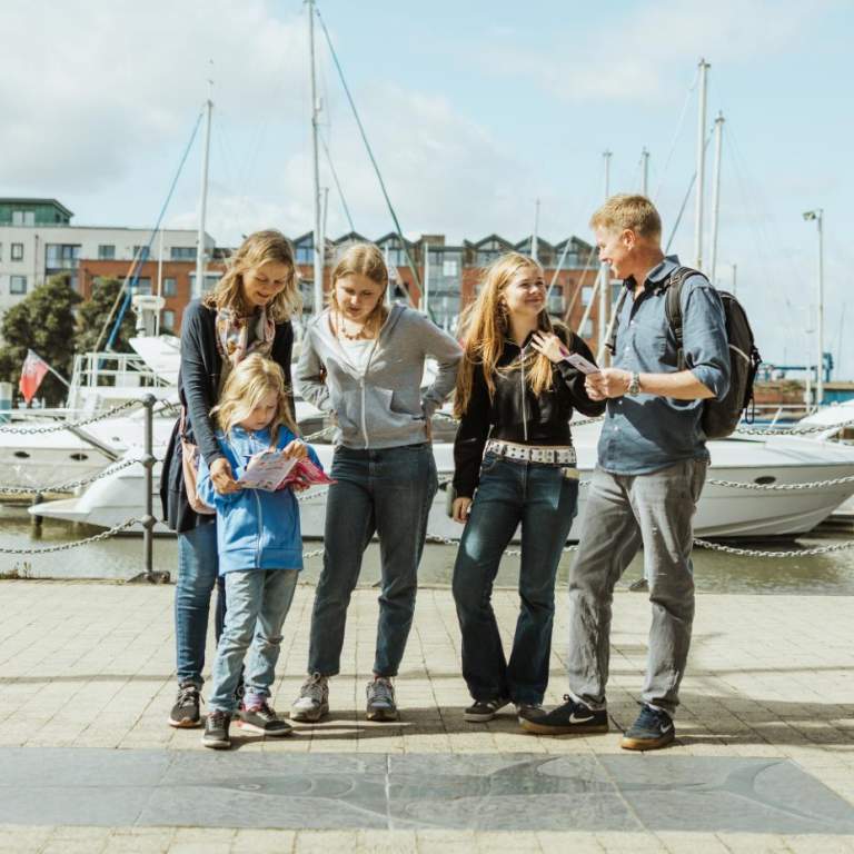A group of people standing beside a stone fish carving on Hull’s Fish Trail near the marina, holding trail maps with boats and water in the background.