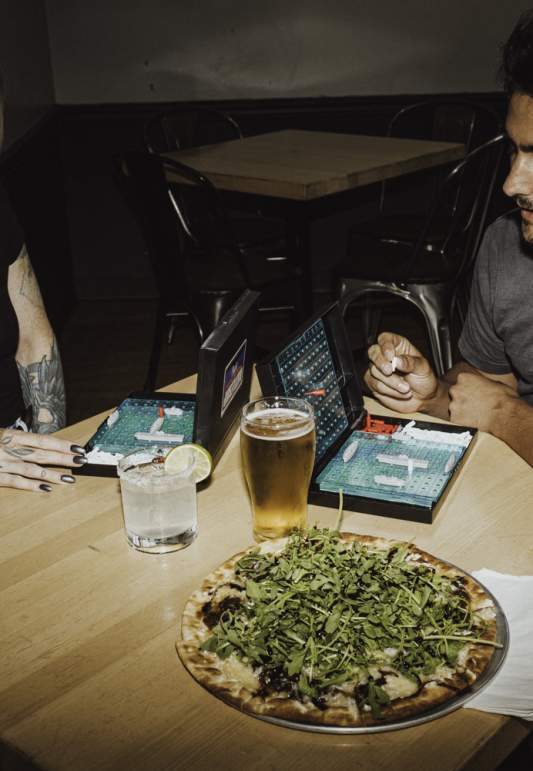 Two people play a tabletop game at a restaurant table with pizza and drinks in front of them.