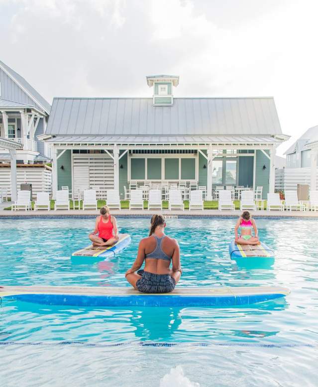A woman with crossed legs sits atop a surf board floating in a pool, back to the camera. Four women, also with crossed legs, sit atop surf boards facing the instructor.