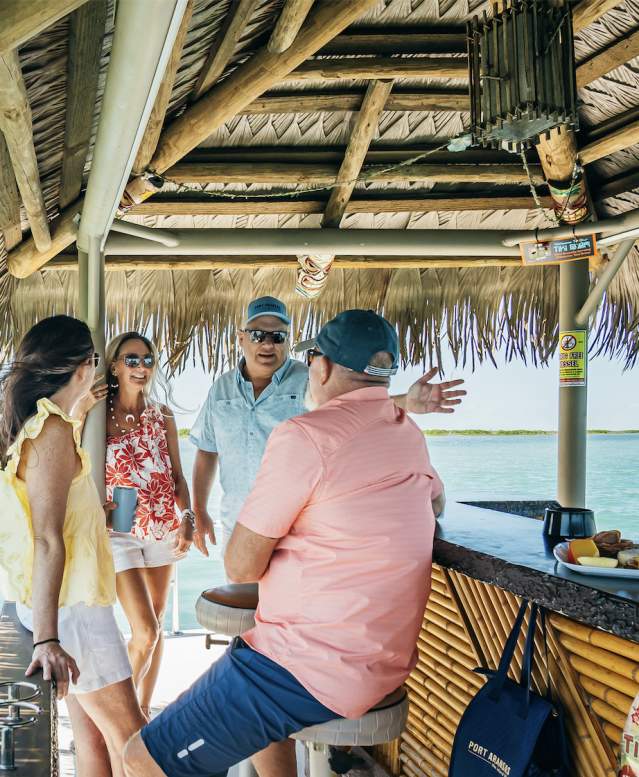 A group of four enjoy a day on a tiki boat. They converse in the foreground with a palapa roof overhead and a view of the Lydia Ann Lighthouse in the background.