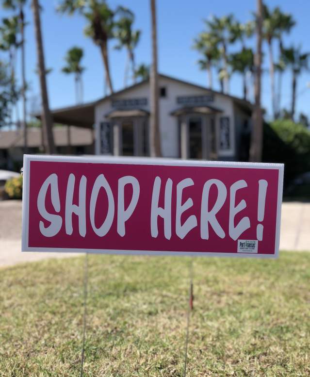 A rectangular pink sign reading "Shop Here!" takes up the middle of the photo. A building surrounded by palm trees is visible in the background.