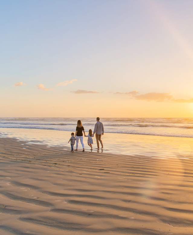 A family of four walks on the beach away from the camera towards the left. The sun is rising to the right.