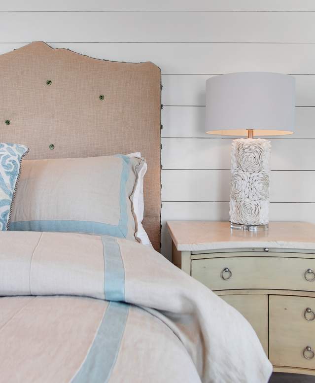 Image of a tan bed and nightstand against a white shiplap wall. On top of the nightstand is a shell lamp.