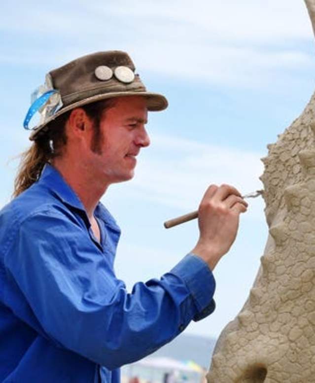 Sand sculptor in a hat with a feather and buttons sculpting the head of a dragon in sand.