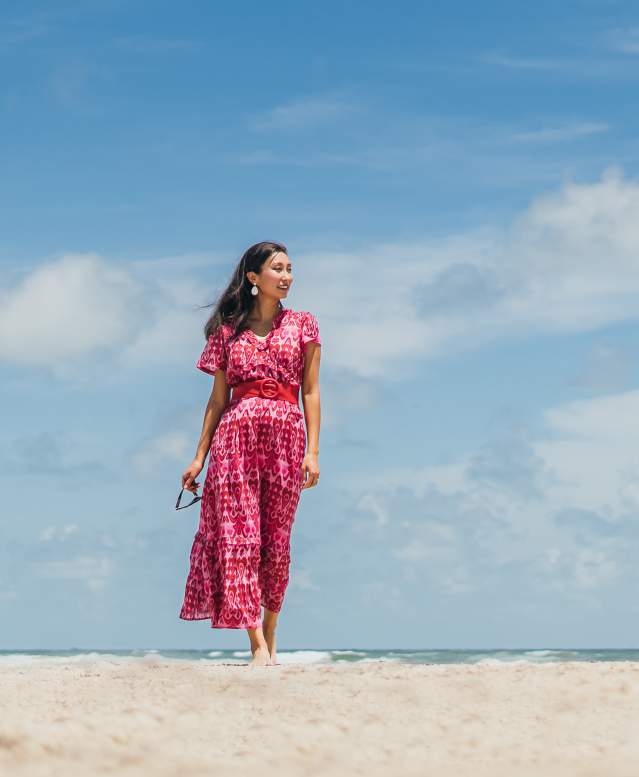 A woman in a pink and red dress walks along the beach