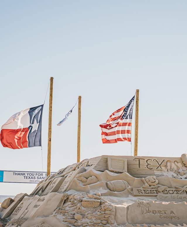 Three flag poles with the US flag, Texas flag, and a Texas SandFest flag fly on poles behind a large sand mountain carved with sponsor logos