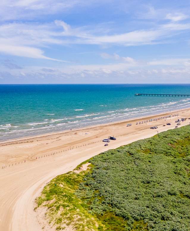 Drone image of a beach that shows the water, beach, and sand dunes from very high. Down the beach, a few cars and a pier are visible.